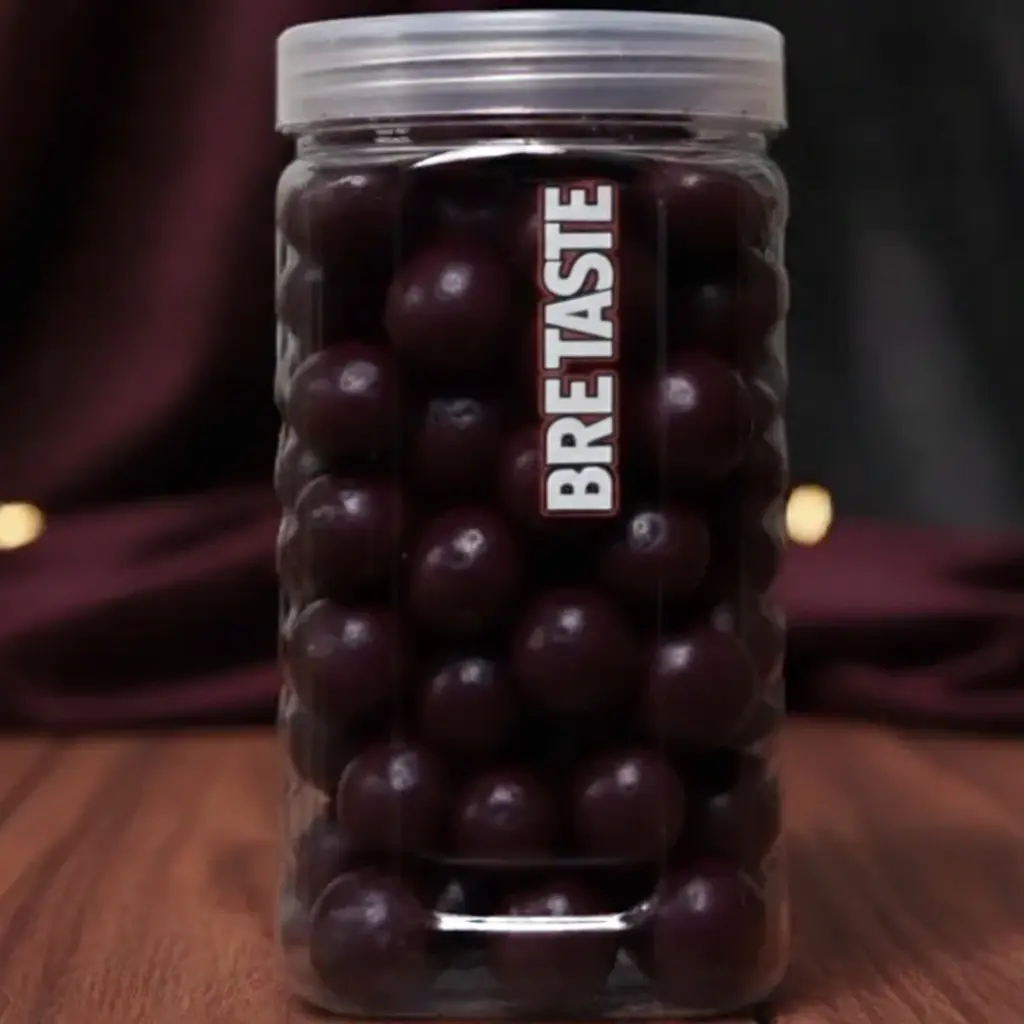 A tall Brektaste jar with a red lid, filled with dark purple, round Jamun Gatagat gola candies, sitting on a wooden table.